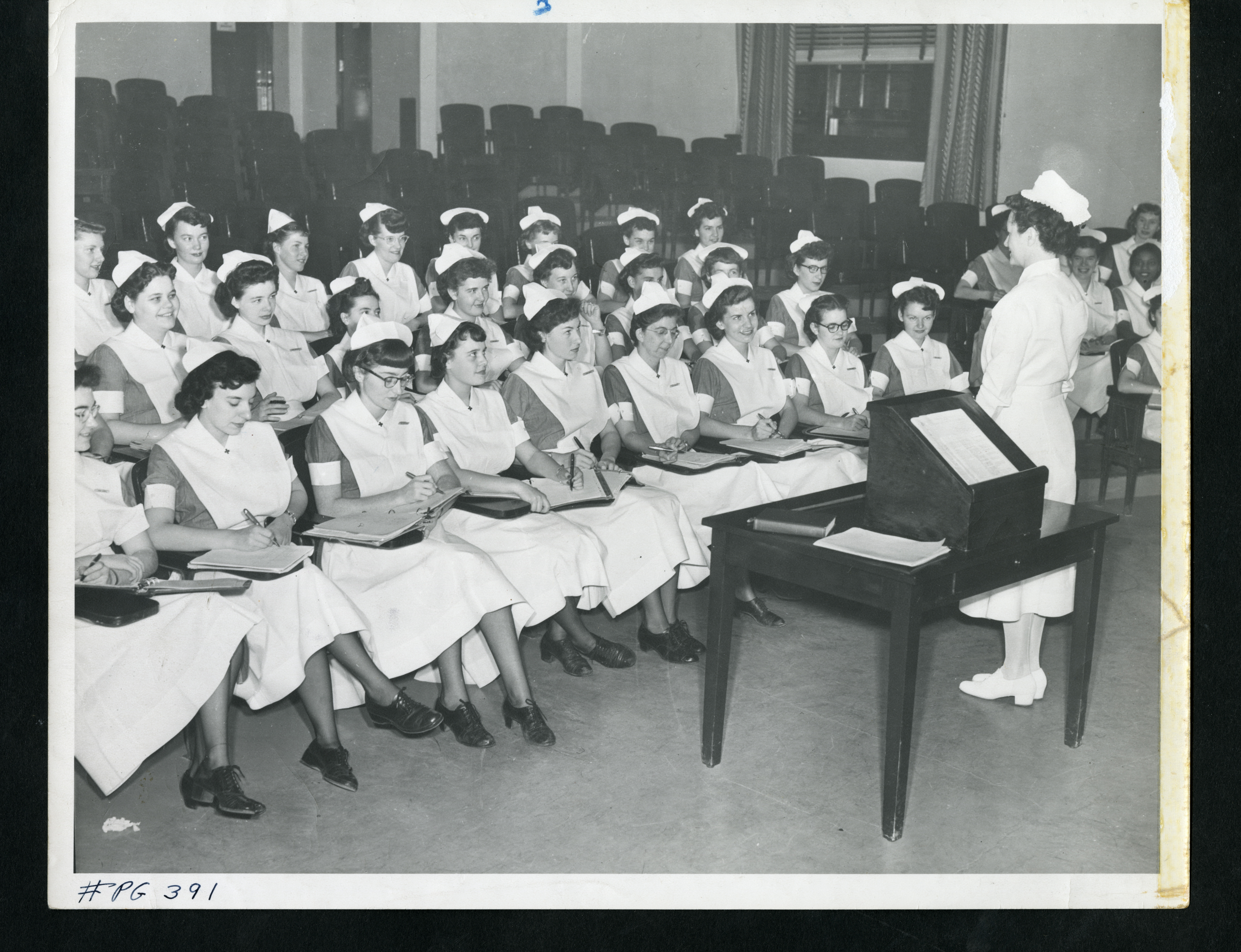 A black and white image of a group of women in nurse uniforms seated in a lecture hall. Another woman in a nursing uniform stands in front of them next to a lectern them.