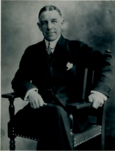 A black and white photograph of Edward Bok, seated with his legs crossed.