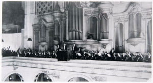 A black and white photograph of the Philadelphia Orchestra led by Leopold Stokowski. The orchestra is on a balcony seated in front of a large pipe organ at Wanamaker's Department Store.