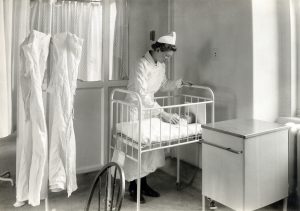 A black and photograph of a nurse in uniform standing over an infant's crib in a hospital room