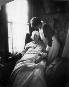 a black and white photograph of a uniformed nurse standing over a seated elderly woman in the elderly woman's home.