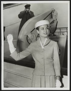 a black and white photograph of Grace Kelly waving. Behind her, a photographer takes a photograph.