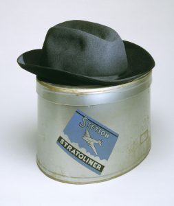 a color photograph of a grey fedora-style hat on a metal hat box with a lable. Text on lable: Stetson Stratoliner