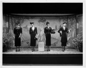 a black and white photograph of four mannequins dressed in black dresses in a display window of a department store.