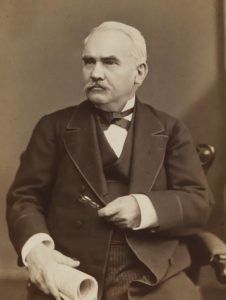 Photogrpah of Morton McMichael. The image is sepia toned. McMichael is an older white man wearing a three piece suit, seated and looking slightly to his right. He holds a rolled up paper in one hand and his pipe in the other, which is across his chest.