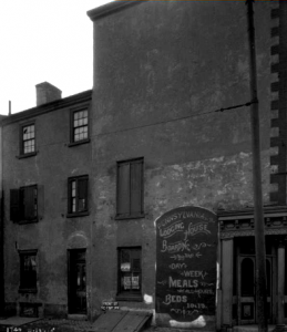 a black and white photograph of a building with an advertisement for boarding accomodations painted on the front. Text reads "Pennsylvania Lodging House, Boarding by the Day or Week, Meals served at all hours, Beds 10 & 15"