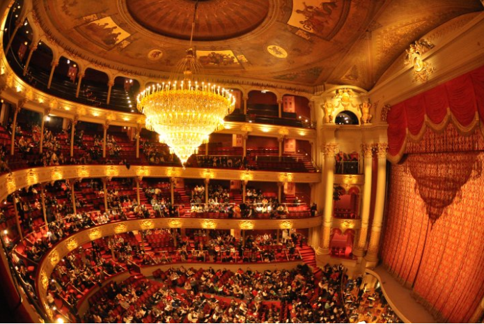 A color photograph of the interior of the Academy of Music showing stage, proscenium, and audience seated under a large chandelier.