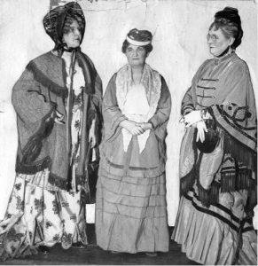 a black and white photograph of three women dressed in costumes reflecting the fashions of 1877.