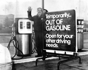 a black and white photograph of a gas station attendant standing next to a gas pump and a sign. Text on sign: "Temporarily out of gasoline. Open for your other driving needs"