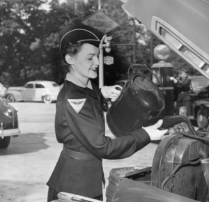 a black and white photograph of a woman pouring radiator fluid into a car. She is wearing a work uniform, white gloves, and a hat. 