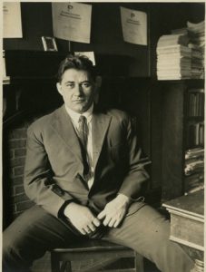 Black and white photograph of Michael Dorizas sitting, wide legged, behind his desk at the university of pennsylvania. He is middle aged with dark hair and wears a full suit with tie. 