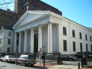 Color photograph of st. george's greek orthodox cathedral. The shot is taken from across the street and shows a large white stone building with two floors of windows along the sides. There are greek style columns in the front and a small white cross at the top of the arched roof.