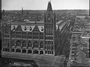 A black and white photograph of Broad Street Station, a gothic revival building with a church-like steeple. Attached to the rear is a large train shed and a viaduct that stretches into the distance. Residential and commercial development runs along either side of the viaduct.