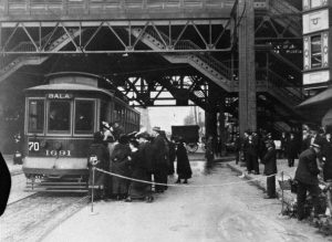 A black and white photograph of a group of people waiting to board a street car. The street car is stopped under the elevated tracks of the Market Frankford Line