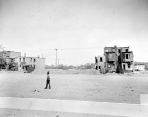 A black and white photograph of a young man walking on a desolate stretch of sidewalk. Behind him two small clusters of dilapidated row homes stand. The rest of the block has been razed.