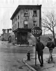 A black and white photograph of two men standing on a street corner. One leans on a stop sign. Across the street is a boarded up, graffitied row house, a dead tree, and scattered litter and debris.