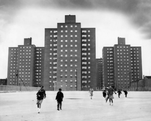 a black and white photograph of a small group of children playing in an empty, paved lot. Behind them, a group of three identical high rise housing project towers looms somewhat ominously.