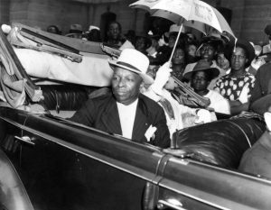 Father Divine, an African-American man wearing a pinstripe suit and hat, sits in an automobile. He is surrounded by people who are all hoping to catch a glimpse of him during his visit to Philadelphia. His wife Penninah is seated next to him and carries a parasol.