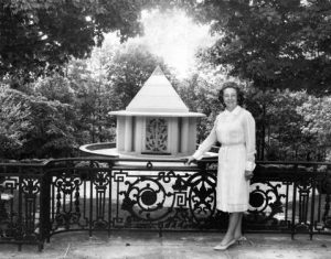 Mother Divine is shown next to a pyramid-shaped memorial at Father Divine's burial site. She is wearing a white dress and has her hand resting on a fence that serves as a barrier between her and the memorial.