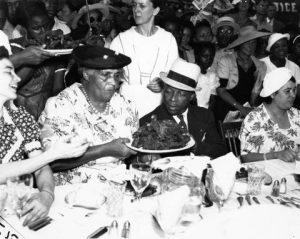 Father Divine, wearing a black suit and white hat, sits next to his wife Penninah, who wears a floral-print dress. They are seated at a large table at Shibe Baseball Park in Philadelphia. Penninah passes a plate of food to Father Divine while other guests enjoy the meal in the background.