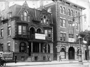 A black and white photograph of two buildings on a city street in 1942. One is three stories and the other is four stories. A few people walk past in the foreground.