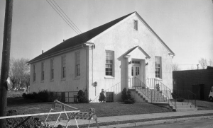 A black and white photograph of a small, single-story church. It is painted white and is surrounded by a few trees.