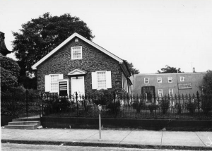 A black and white photograph of a single-story stone meetinghouse. It is surrounded by a black fence and there is a two-story building behind it.