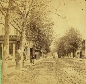 A black and white photograph with a yellow tint that depicts a quiet Main Street. A few buildings are visible in the background, as well as two men standing by a tree and having a conversation. The streets are clear.