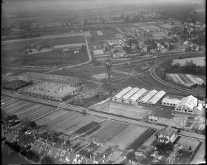 This black and white photograph shows a large factory surrounded by houses and open plains. A water tower sits in the middle of the property and several can production buildings sit on both sides.