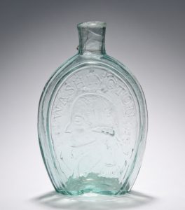 This photograph shows a glass flask made at the Bridgeton Glass Works. A profile of George Washington is engraved on the front and surrounded by text that reads "Washington."