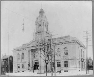 This black and white photograph shows a large courthouse with large glass windows and a central tower. It is surrounded by utlity poles and lights.