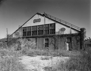 This black and white photograph shows a deteriorated machine production facility. It has a long, narrow roof and several windows are broken. Weeds cover the pavement in the foreground.