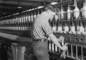 This black and white photograph shows a worker fixing spinning machines in a textile factory. His sleeves are rolled up as he replaces large spools of thread.