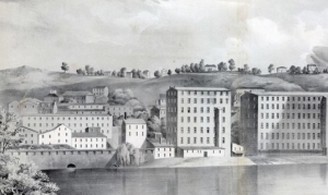 This black and white photograph shows a group of large factories bordering a river. A few houses and trees are visible on hills in the background. One of the factories has an opening in the middle that spills into the river.