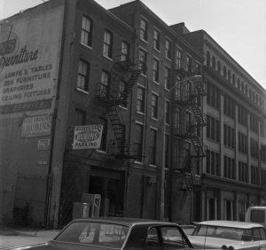 This black and white photograph shows the Wireworks building on Race Street in Philadelphia. An advertisement for a furniture store can be seen on the side of the building and a few 1970s cars sit in the foreground.