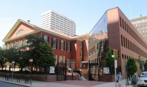 This color photograph shows a large brick building in Center City, Philadelphia. The structure on the left has an older architectural style with two stories and several glass windows, while the structure on the right looks more modern and has a large glass surface facing inward.