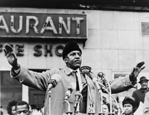 This black and white photograph shows Bayard Rustin, a civil rights activist, as he addresses as crowd in New York City. Rustin's arms are raised and there are several microphones for TV and radio stations on the podium in front of him.