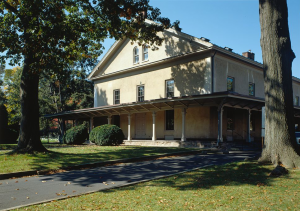 This color photograph depicts the Germantown Friends Meeting House. The building has a tan-yellow paint color, a long secondary roof that runs above the first floor, and a few windows at symmetrical points on all three floors.