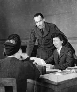 This black and white photograph shows a man and woman in American Friends Service Committee uniforms talking to a woman seated at a table. The uniformed woman writes notes while the uniformed man looks at the camera.