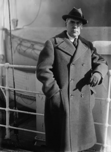 This black and white photograph shows Albert C. Barnes on the deck of a boat. He wears a large winter coat, gloves and hat and leans against the railing. Two smaller boats can be seen in the background.