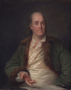 This is a painting of Benjamin Franklin, gesturing towards a piece of paper sitting on a desk next to a pair of bifocals. He is wearing a green, fur-lined jacket.