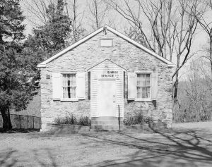 This black and white photograph shows a small stone church resting atop a hill. Several trees and a fence are visible in the background.