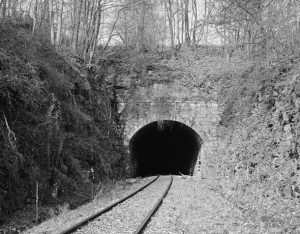 This black and white photograph shows a tunnel connected to a section of railroad track. The area is surrounded by trees and rocks.