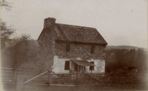 A black and white photograph of a two-story stone home with a chimney; the first story is white washed.