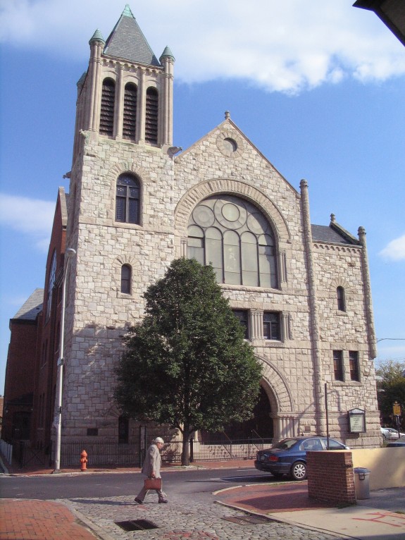 This color photograph shows the Mother Bethel AME Church. A large stained glass window appears near the top and the outer later is comprised of layered stone.