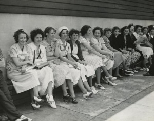 This black and white photograph shows a group of fourteen women seated on a bench outside the Apex Hosiery factory as they prepare for a strike.