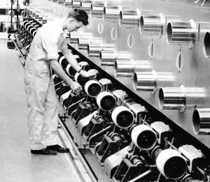 This black and white photograph shows a worker adjusting the threads of a large nylon machine. There are several large round spools close to him leading to a polished steel structure above.