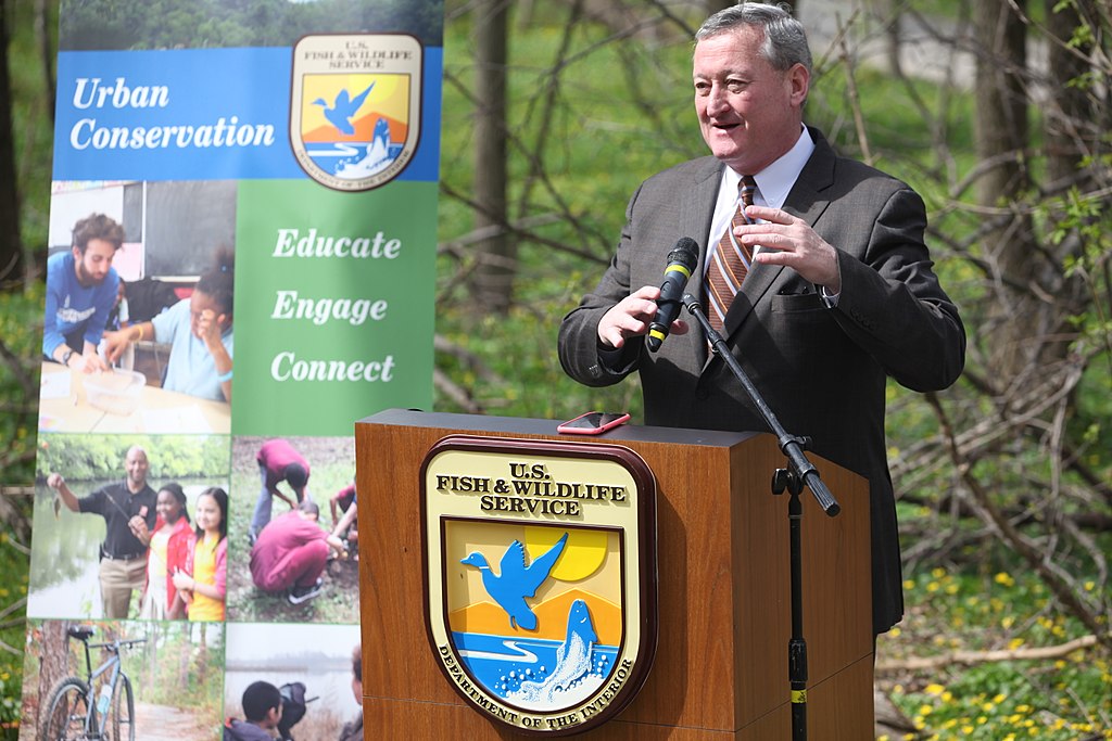 This color photograph shows Jim Kenney standing at a podium labeled "U.S. Fish and Wildlife Service." A poster behind him reads: Urban Conservation - Educate, Engage, Connect.