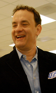 This color photograph shows actor Tom Hanks. He is wearing a blue collared shirt and a black jacket with a label that reads "US."