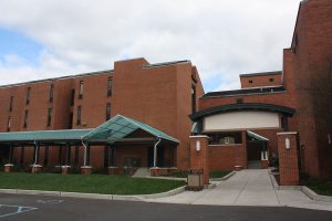 This color photograph shows a large brick building with several windows. A large patch of grass separates the building from its parking lot.
